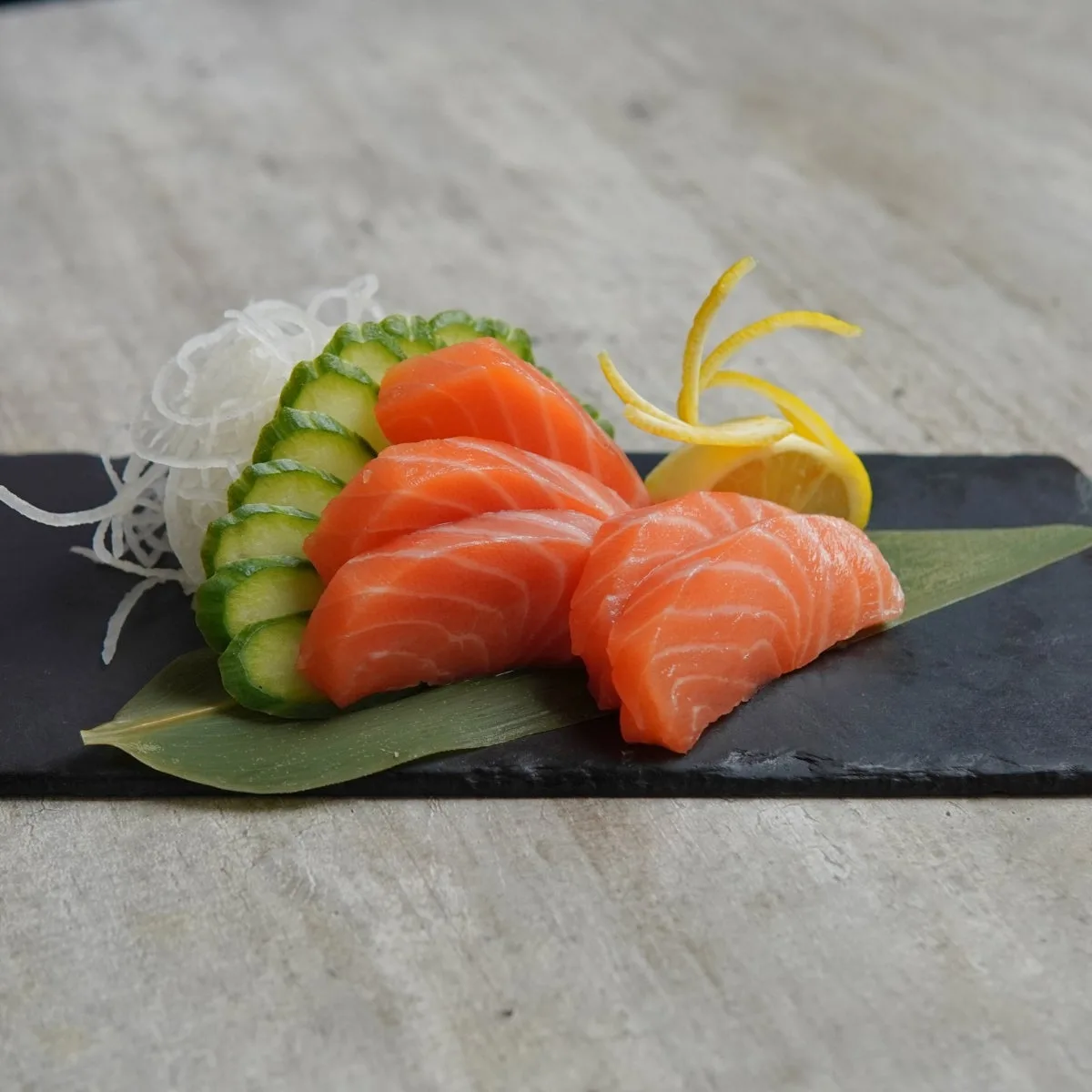 Slices of salmon sashimi with cucumber and lemon on a slate board